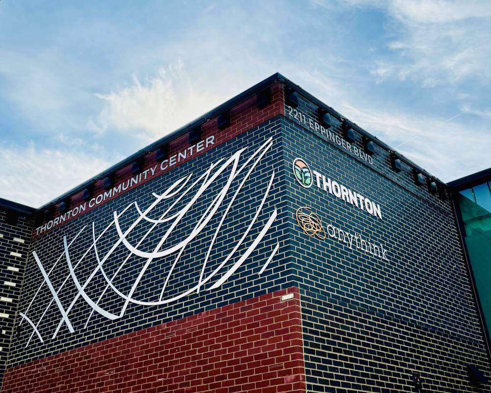 An exterior brick wall of a building. Sign on wall in English reads Thornton Community Center, 2211 Eppinger Blvd. The city of Thornton and Anythink libraries logos are on the wall. 