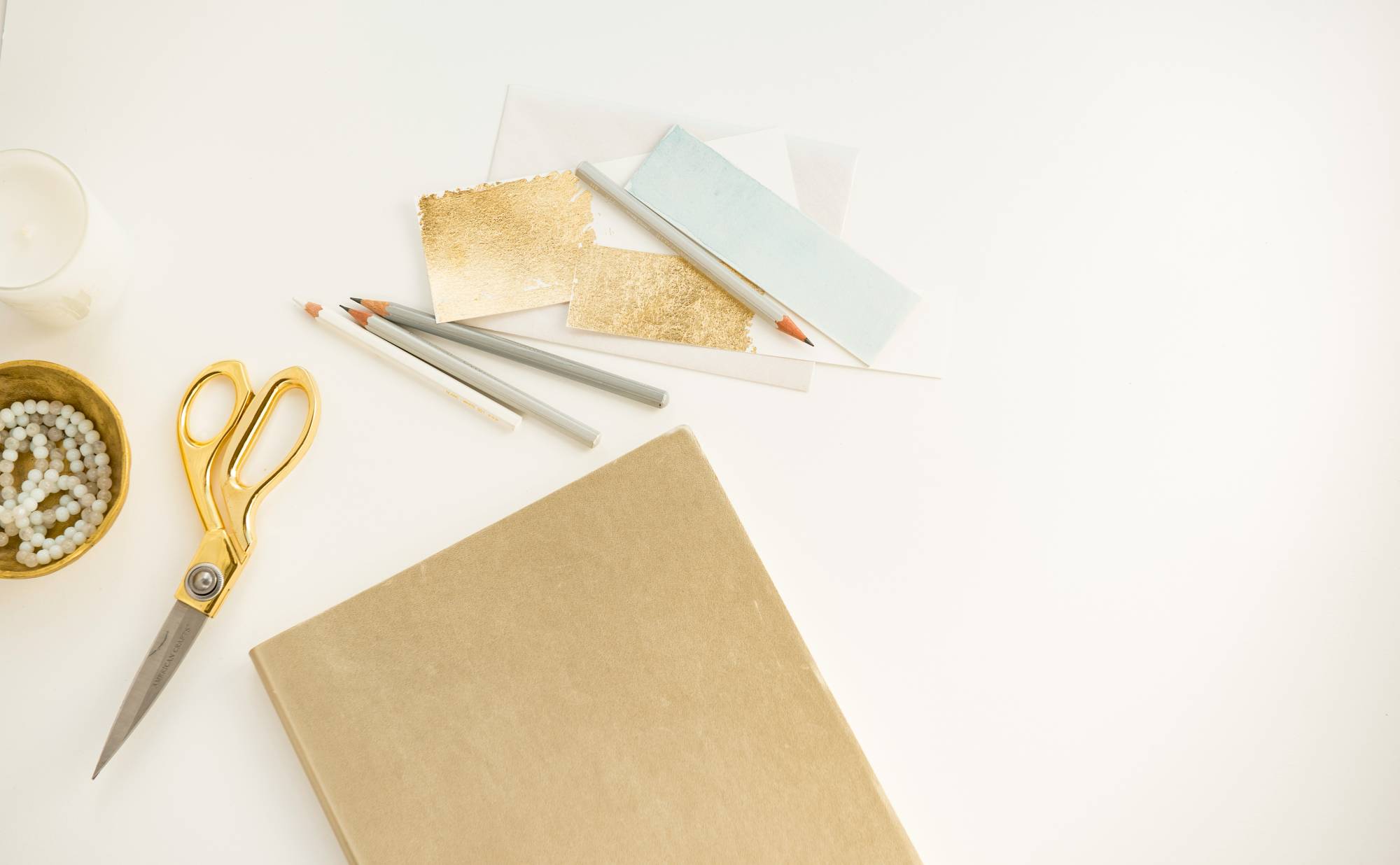 A white tabletop with blank brown paper, gold scissors, pencils and other supplies on it.