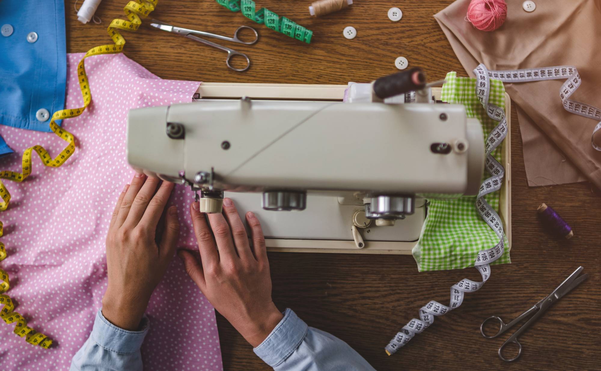 A person's hands using a sewing machine on a tabletop with fabric, ribbon, and sewing tools strewn about. 