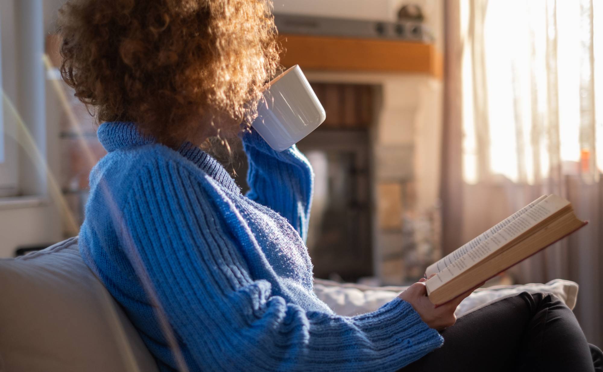 An adult woman with a natural afro hairstyle sitting on a couch with an open book in one hand, and sipping from a mug. 