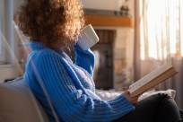 An adult woman with a natural afro hairstyle sitting on a couch with an open book in one hand, and sipping from a mug. 