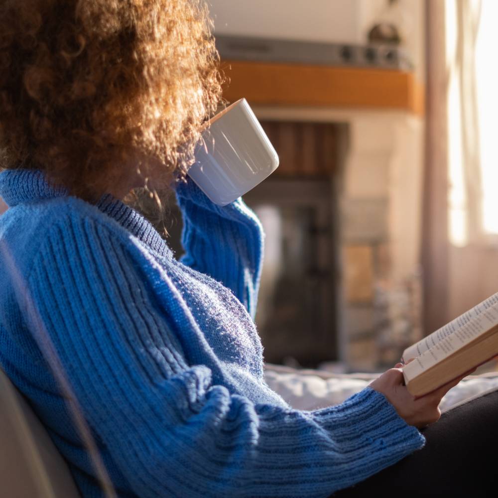 An adult woman with a natural afro hairstyle sitting on a couch with an open book in one hand, and sipping from a mug. 